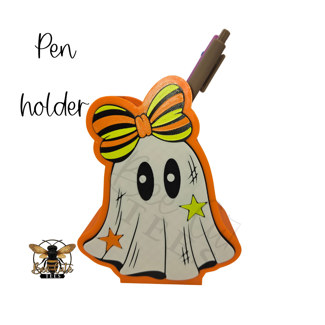 A Halloween-themed ghost-shaped pen holder with an orange border, a striped orange and yellow bow, and star details, shown on a white background with a pen inserted. The text 'Pen holder' appears on the left, along with a bee logo.