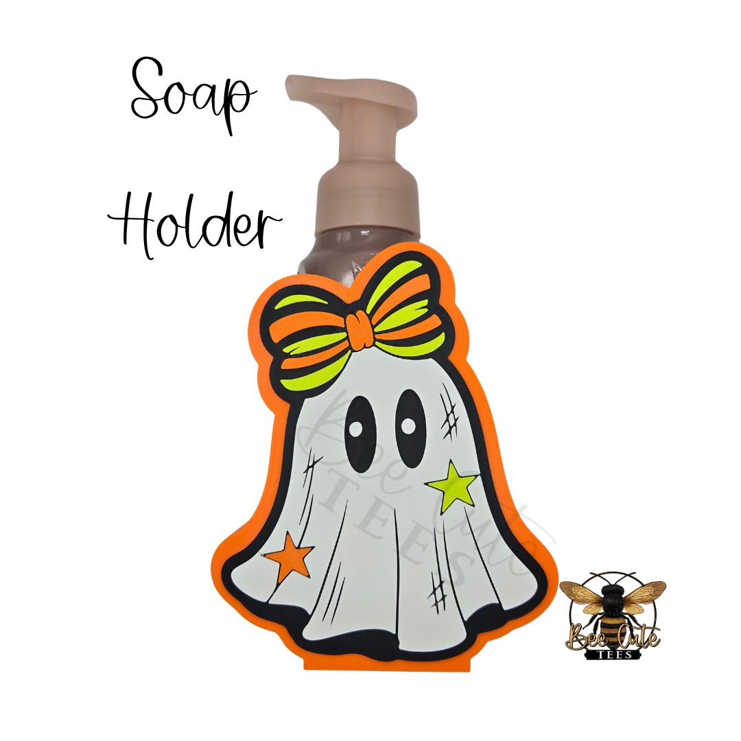 A Halloween-themed soap dispenser with a ghost-shaped protective cover featuring an orange bow, orange stars, and a black outline, shown on a white background with 'Soap Holder' text and a bee logo.