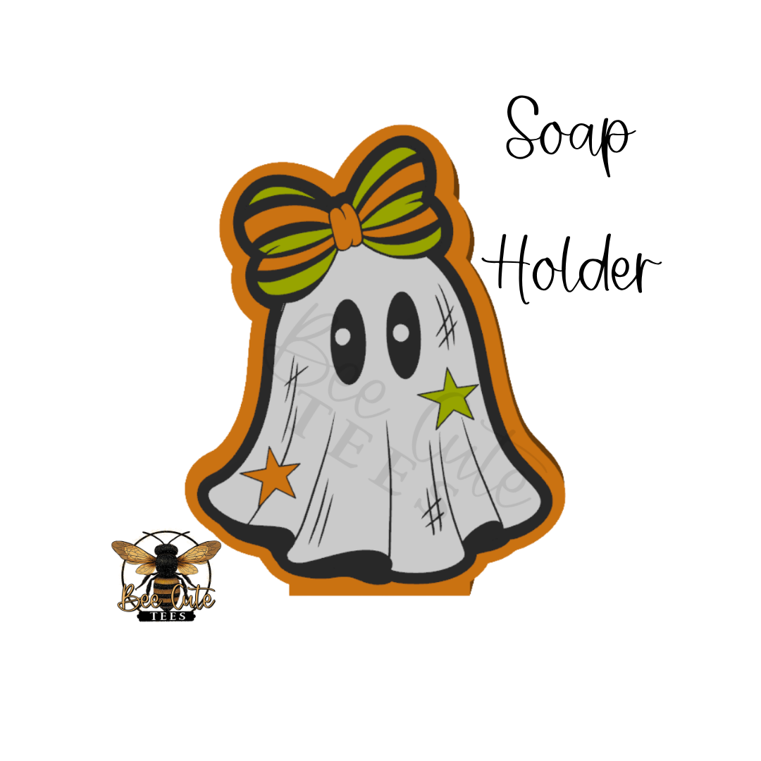 A Halloween-themed ghost-shaped soap holder or plaque with a white ghost body, black facial features and outline, an orange border, a green-and-orange bow, and orange and green stars. The words 'Soap Holder' appear to the right, and a small bee logo  at lower left on a white background.