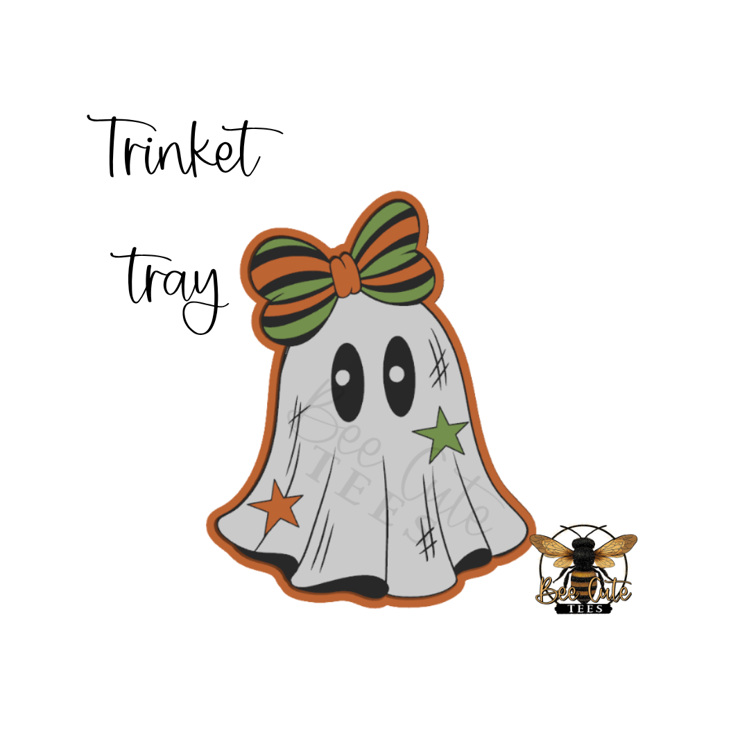 A Halloween-themed graphic showing a ghost-shaped decoration with a green and orange striped bow, black eyes, scratch marks, an orange star, and a green star, with the words 'Trinket tray' above it and a small bee logo at bottom right.