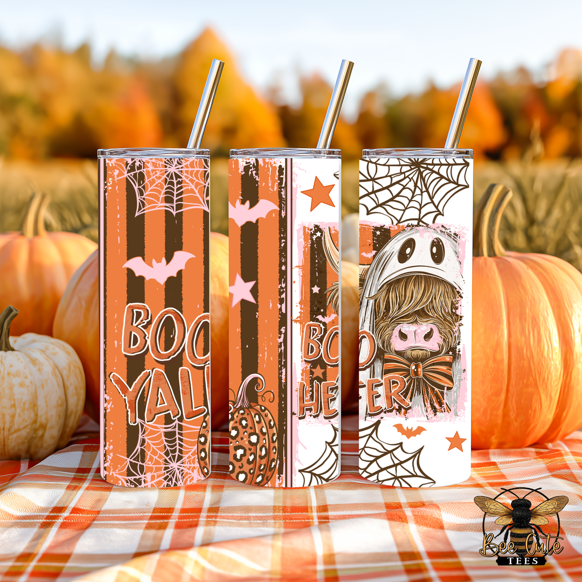 Three Halloween-themed stainless steel tumblers with metal straws, standing on an orange plaid blanket outdoors among pumpkins. The tumblers feature spooky designs including 'BOOYAL' text, bats, pumpkins, spiders, and a cartoon character in a witch hat.