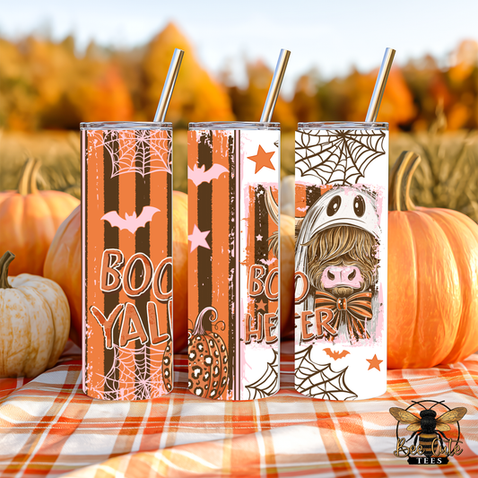 Three Halloween-themed stainless steel tumblers with metal straws, standing on an orange plaid blanket outdoors among pumpkins. The tumblers feature spooky designs including 'BOOYAL' text, bats, pumpkins, spiders, and a cartoon character in a witch hat.