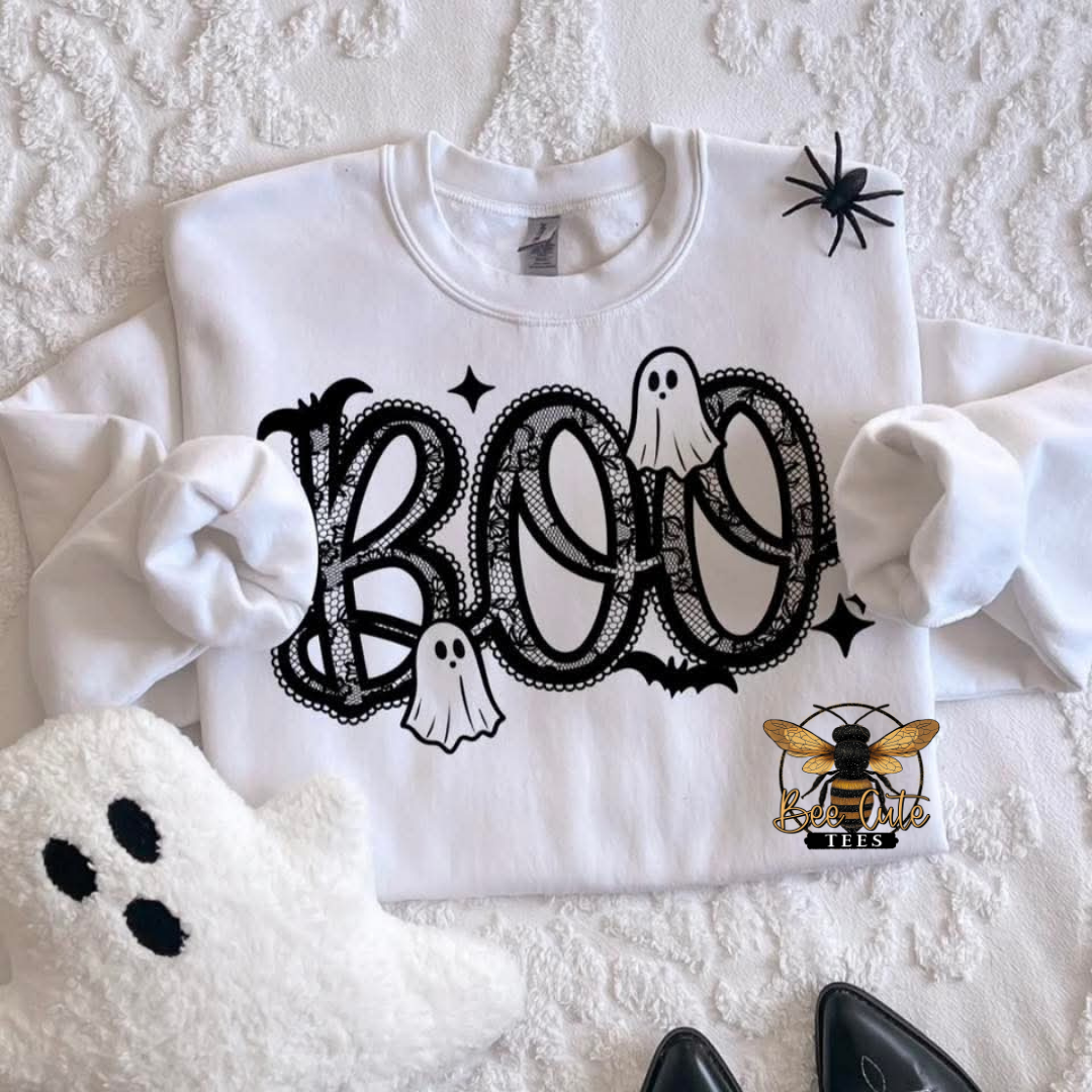 A white crewneck sweatshirt with a black 'BOO' Halloween graphic, featuring ghost and bat details, laid flat on a textured white blanket with a spider, plush ghost, and shoes as props.