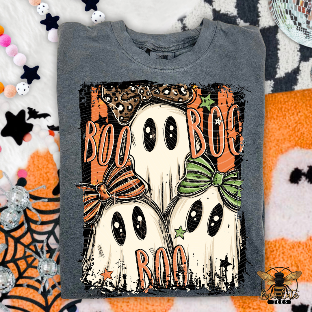 A folded gray t-shirt with a Halloween graphic featuring three ghosts wearing orange and green bows, surrounded by the word 'BOO' in orange. The shirt is laid flat on a white and orange patterned background with Halloween-themed props.
