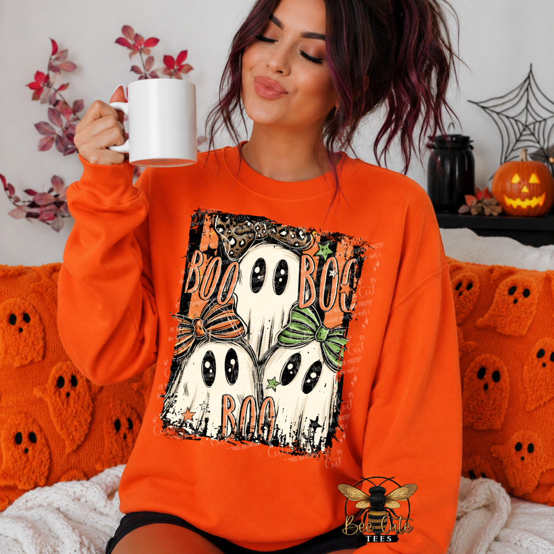 A woman wearing an orange sweatshirt with a Halloween graphic of three ghosts and the words 'BOO BOO BOO' sits on an orange knit couch holding a white mug.