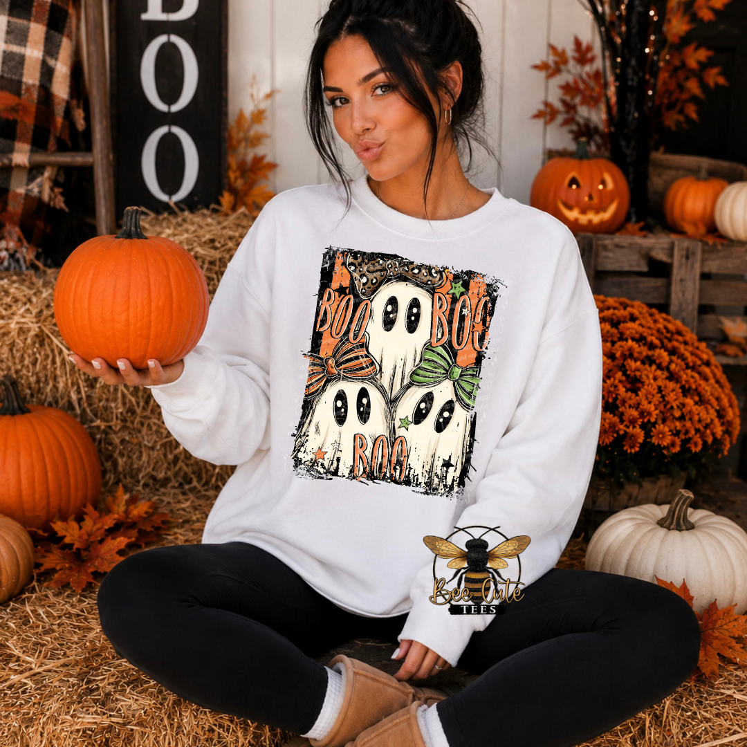A woman sits cross-legged on a bed of hay in a rustic fall-themed setting, wearing a white crewneck sweatshirt with a Halloween graphic of three ghosts and pumpkins. She holds a small orange pumpkin in her hand.