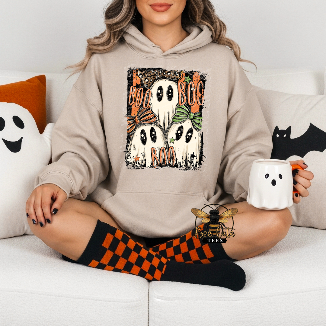 A woman sits cross-legged on a white couch wearing a beige hoodie with a 'Boo Boo Boo' ghost graphic, holding a white mug shaped like a ghost. She is wearing black and orange checkered socks and has Halloween-themed pillows on the couch.