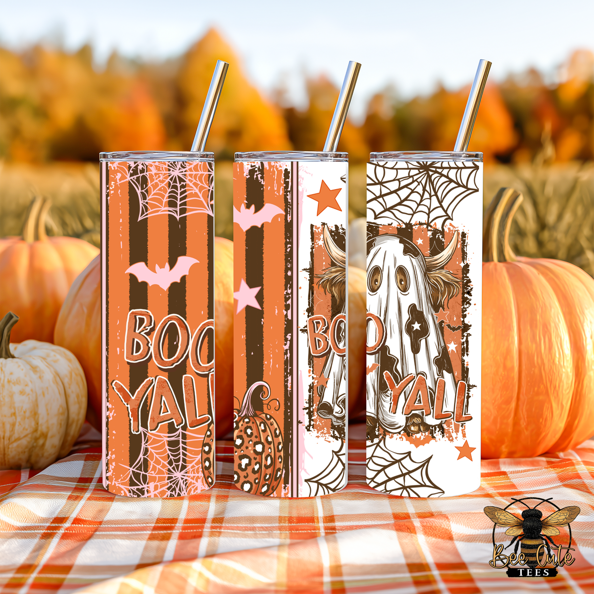 Three Halloween-themed insulated tumblers with metal straws, decorated with spooky graphics like ghosts, pumpkins, bats, and the word 'BOOYAL', displayed on an orange plaid blanket with pumpkins and an autumn outdoor background.