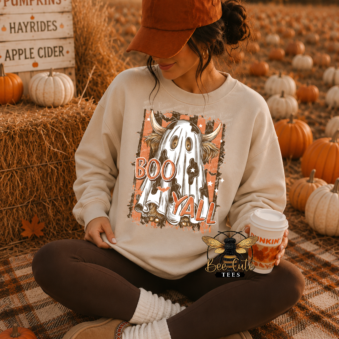 A woman sitting cross-legged in a fall-themed pumpkin field wears a cream sweatshirt with a 'BOO Y'all' ghost graphic, brown leggings, and an orange cap, holding a pumpkin-themed coffee cup.