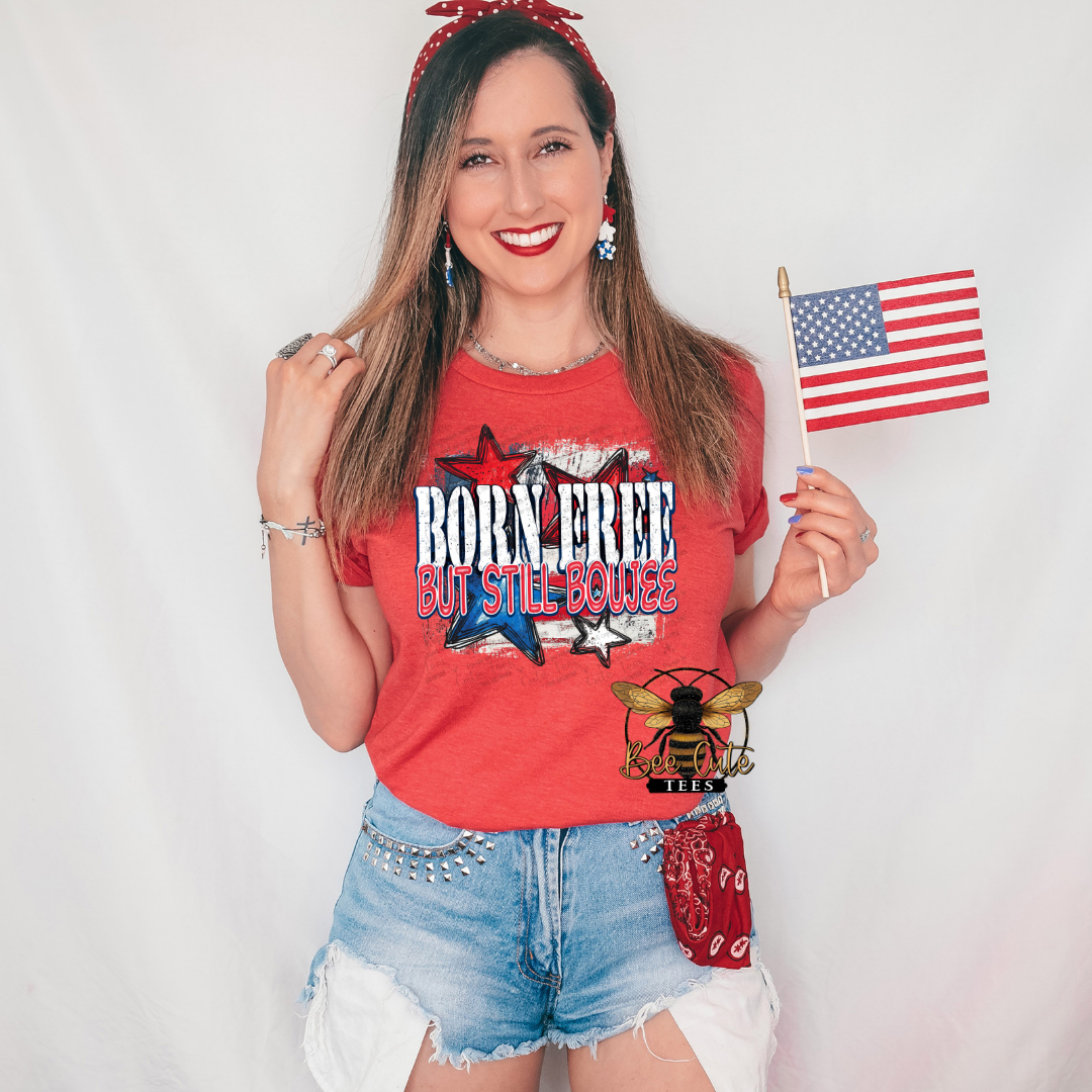 Woman wearing a heather red graphic tee featuring the phrase “Born Free But Still Boujee” with distressed patriotic stars and red, white, and blue accents. Styled with distressed denim shorts, patriotic earrings, a red bandana headband and pocket accessory, and holding an American flag for a fun, festive Americana-inspired look.
