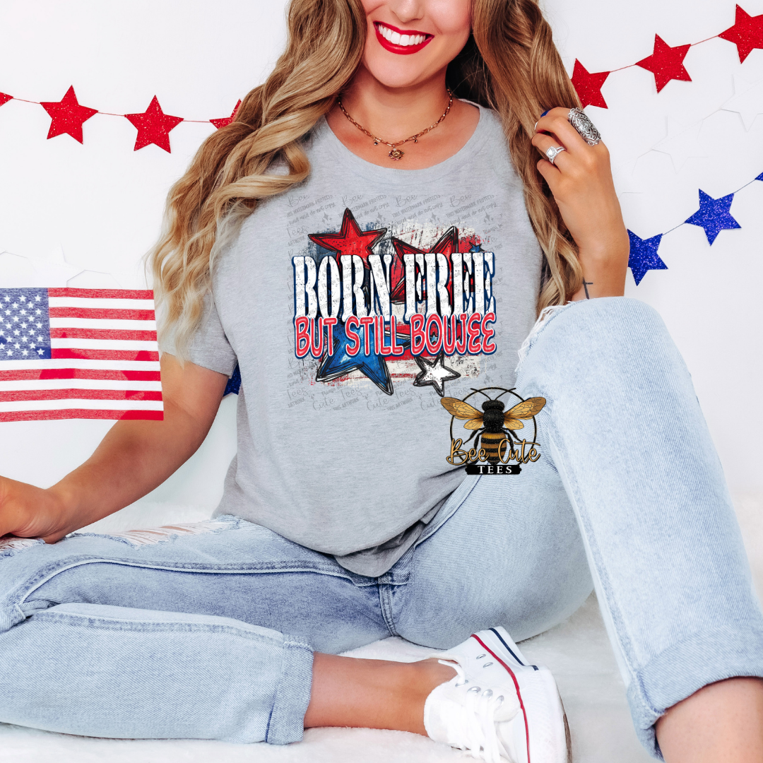 A person wearing a gray t-shirt with a ' Born Free Boujee ' graphic, featuring an eagle, sitting against a backdrop with American flags and stars.