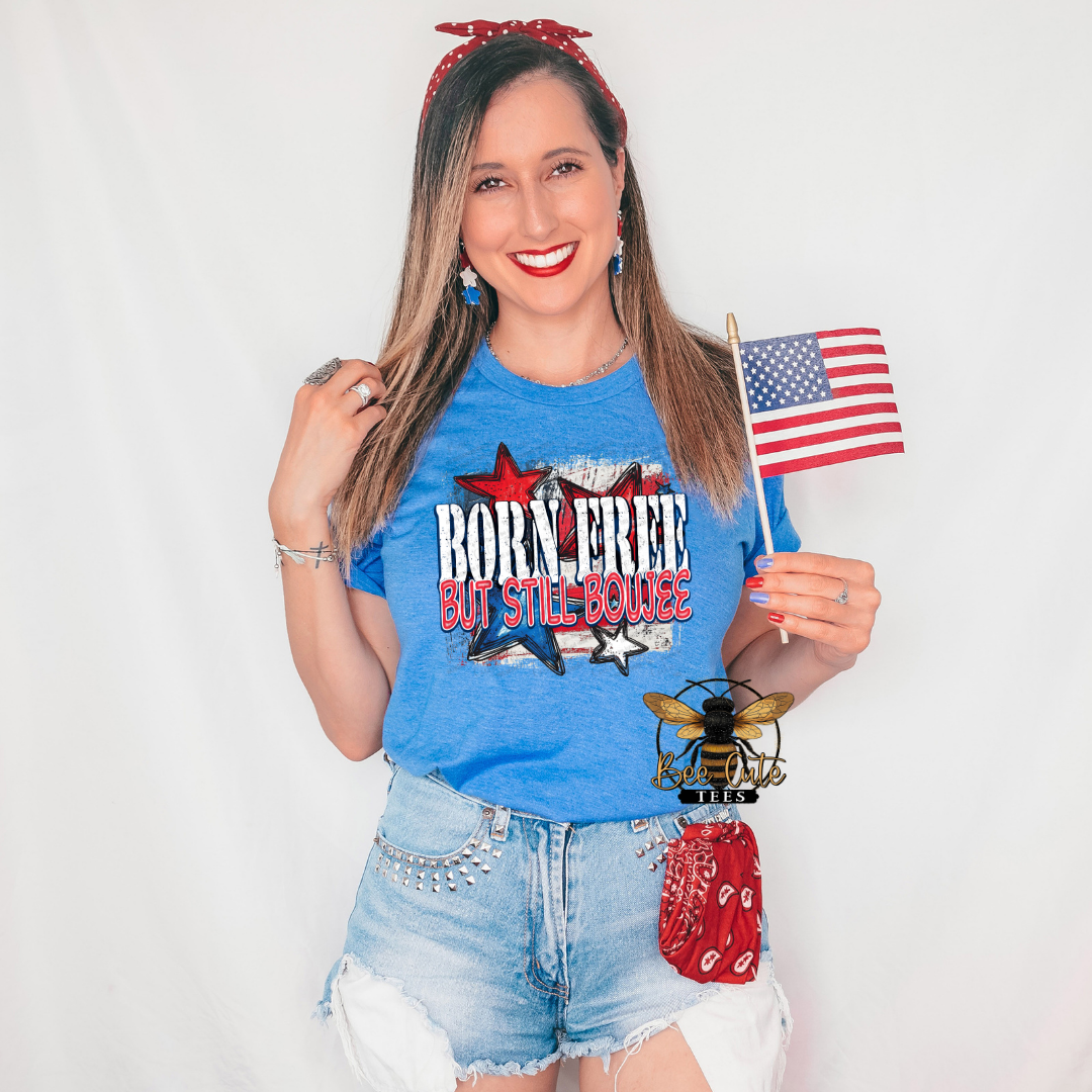 Woman wearing a heather royal blue graphic tee featuring the phrase “Born Free But Still Boujee” with distressed patriotic stars and red, white, and blue accents. Styled with distressed denim shorts, patriotic earrings, a red bandana accessory, and holding an American flag for a fun, sassy Americana-inspired look.
