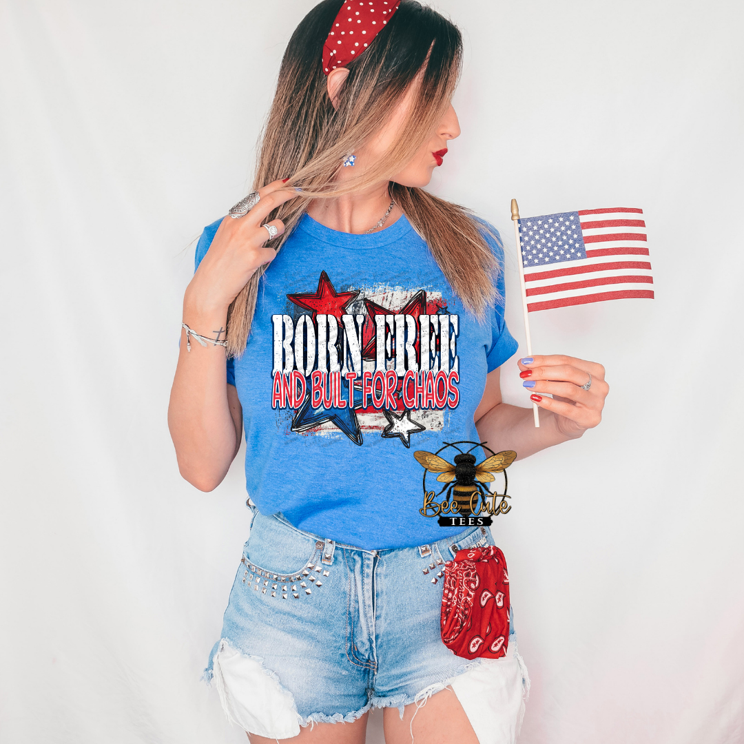 Woman wearing a heather royal blue graphic tee featuring the phrase “Born Free and Built for Chaos” with distressed red, white, and blue stars and patriotic accents. Styled with distressed denim shorts, a red bandana headband, patriotic earrings, and holding an American flag for a bold, fun Americana-inspired look.
