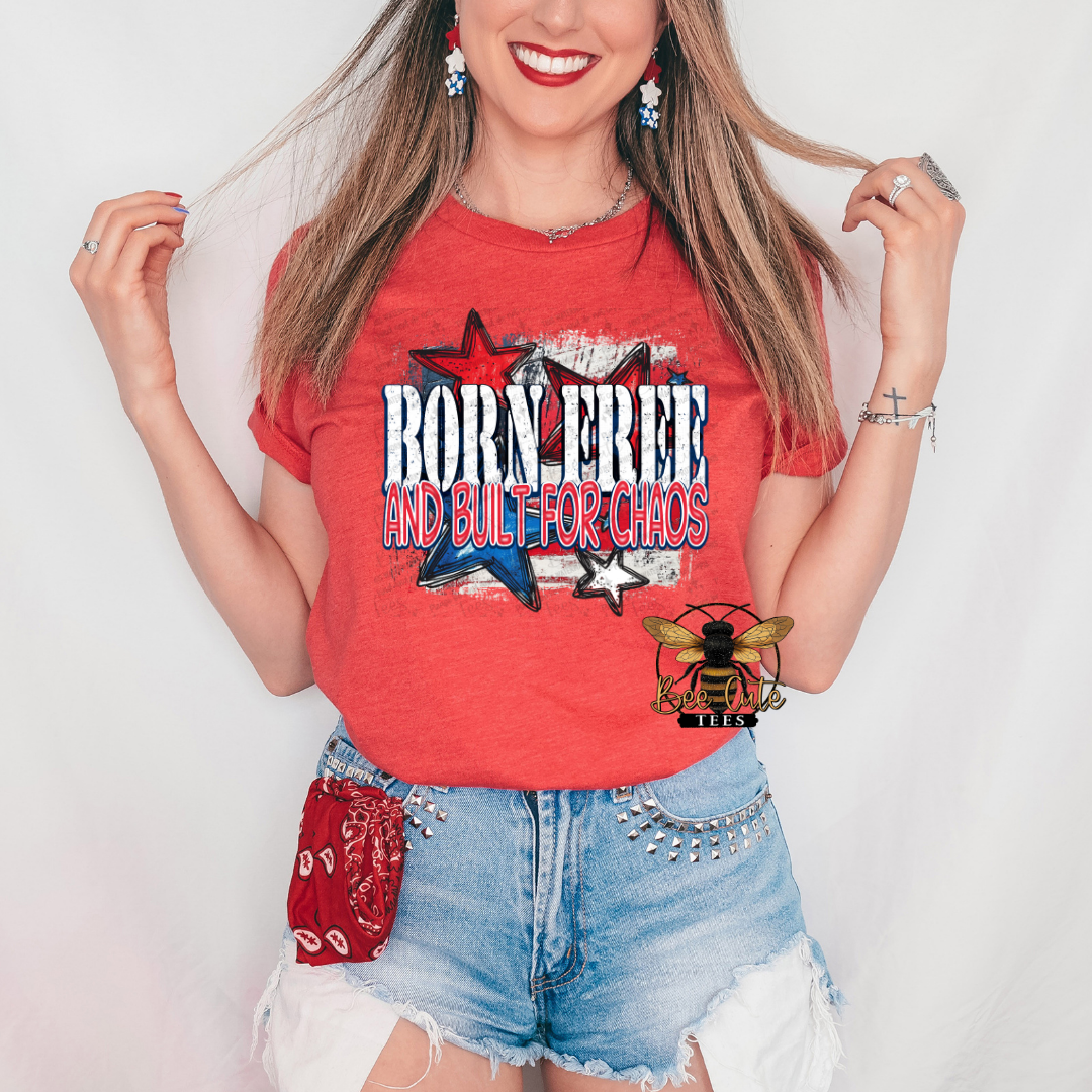 Woman wearing a heather red graphic tee featuring the phrase “Born Free and Built for Chaos” in distressed patriotic lettering with red, white, and blue stars over a weathered American flag background. Styled with distressed denim shorts, patriotic earrings, and a red bandana accessory for a bold, playful Fourth of July look.

