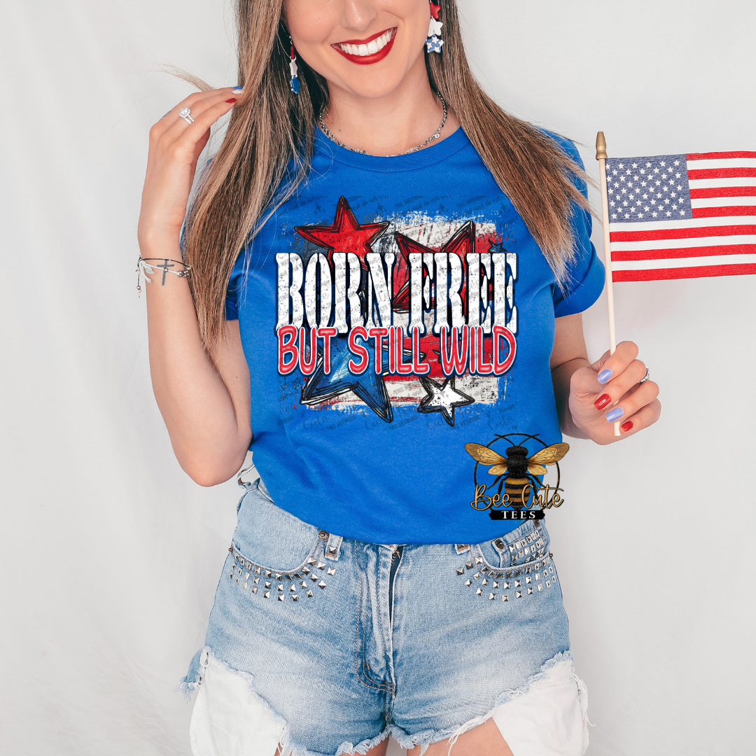 Woman wearing a royal blue graphic tee featuring the phrase “Born Free But Still Wild” in distressed patriotic lettering with red, white, and blue stars layered over a vintage American flag background. Styled with distressed denim shorts, patriotic earrings, and holding a small American flag for a fun and spirited Fourth of July look.
