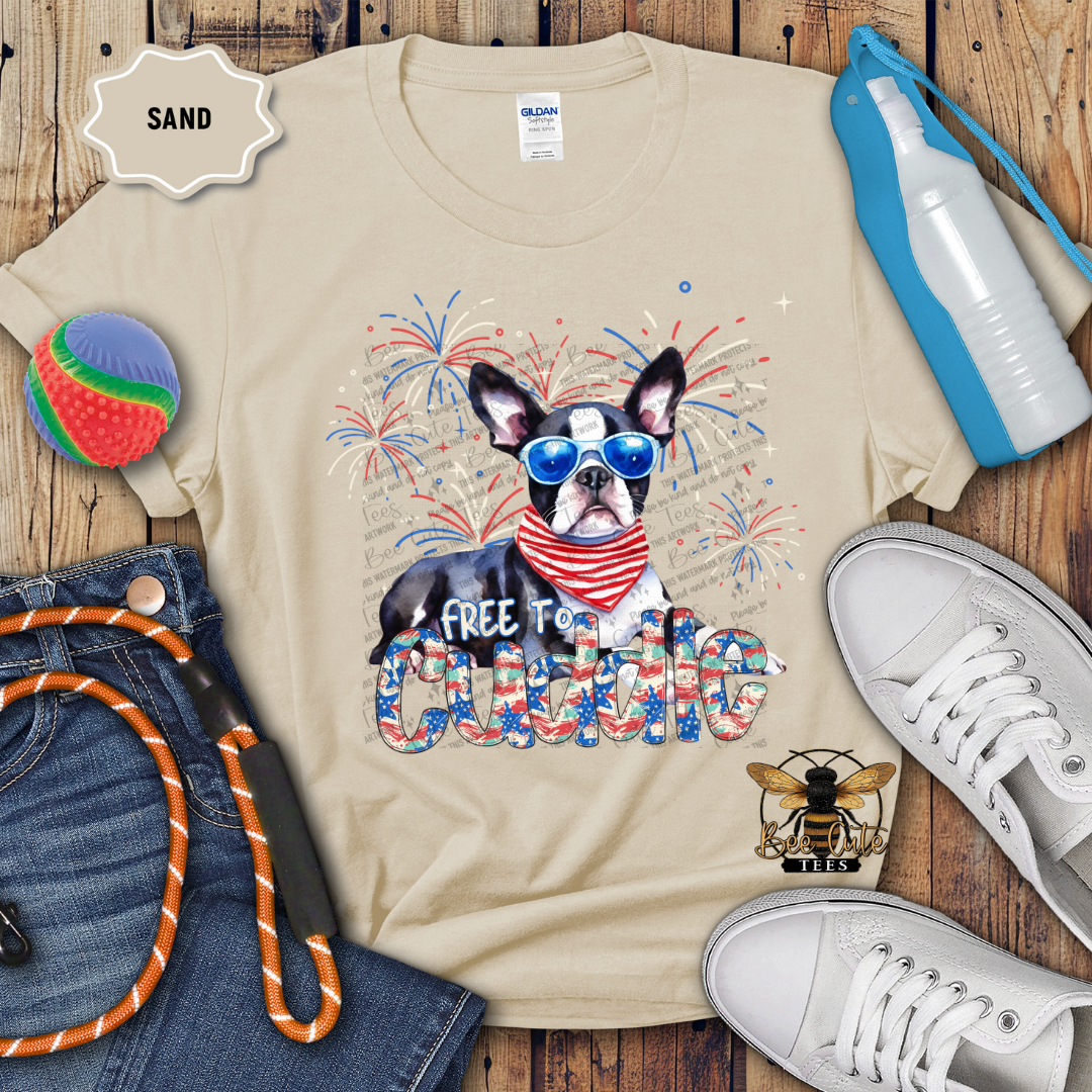 A beige colored t-shirt with a graphic print of a Boston Terrier wearing sunglasses and a neck tie, surrounded by Fourth of July themed decorations such as a patriotic pinwheel, a blue and red water bottle, a rope toy, and a pair of white sneakers. The text 'FREE TO Cuddle' is also visible on the shirt.