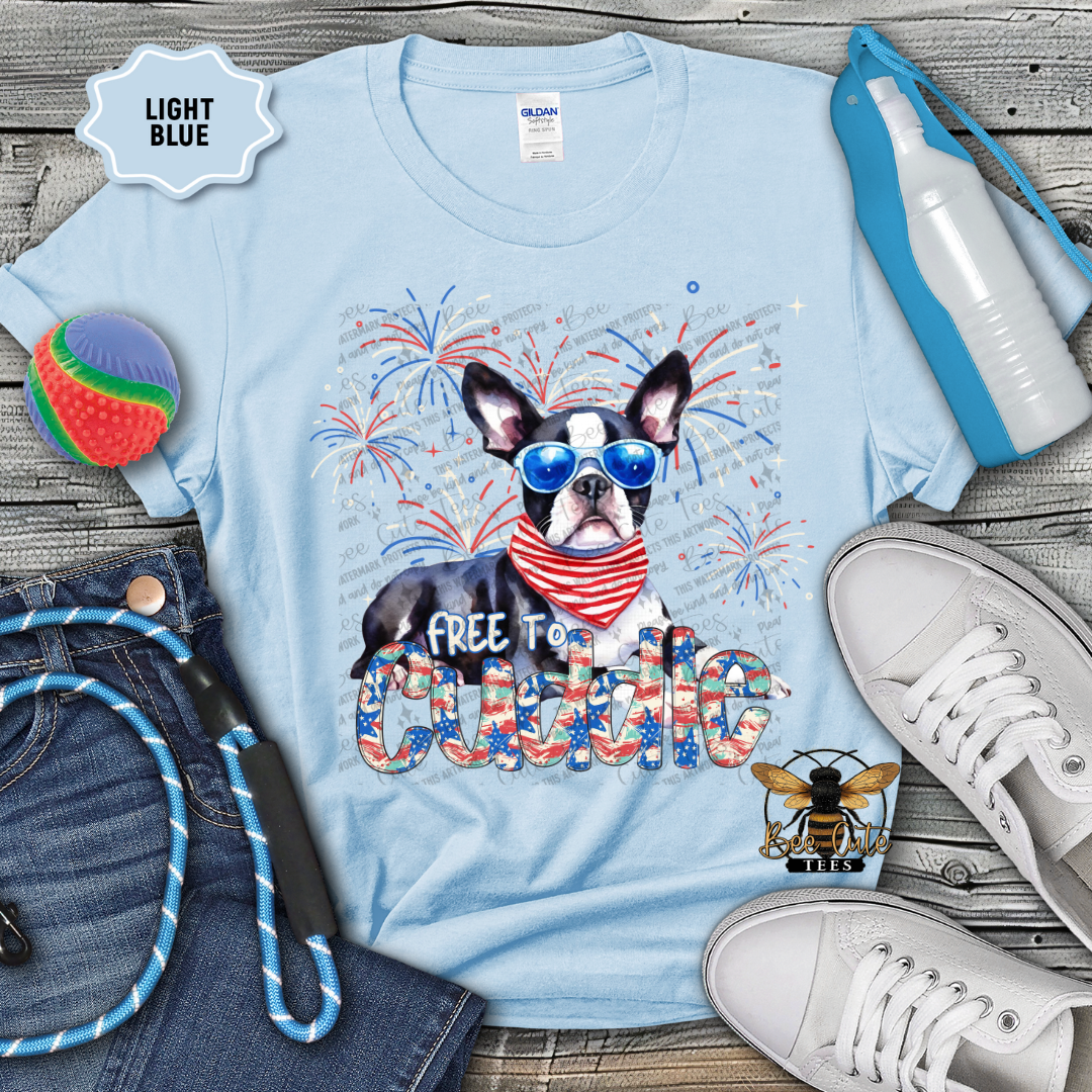 Light blue t-shirt with a dog graphic and 'Free to cuddle' text on a wooden surface with accessories.