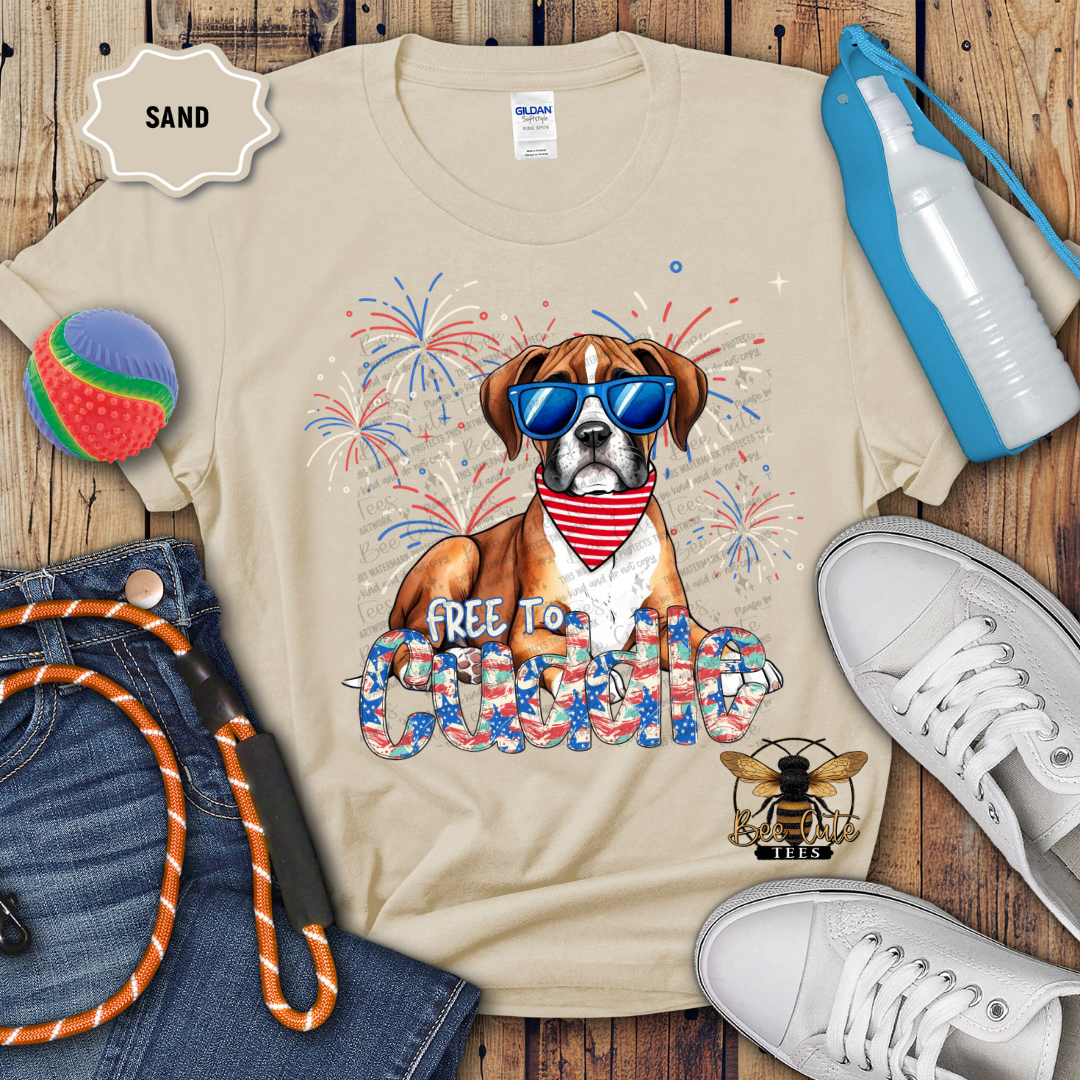 A beige colored t-shirt with a graphic print of a boxer dog wearing sunglasses and a 'FREE TO Cuddle' collar, surrounded by Fourth of July-themed decorations such as a patriotic pinwheel and a flag.
