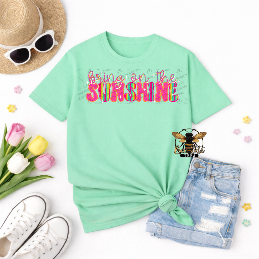 Mint green t-shirt with 'bring on the Sunshine' text, sunglasses, hat, tulips, and shorts on a white background