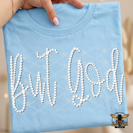 Light blue t-shirt with 'Let Them' text made of faux white pearls, held by a hand.