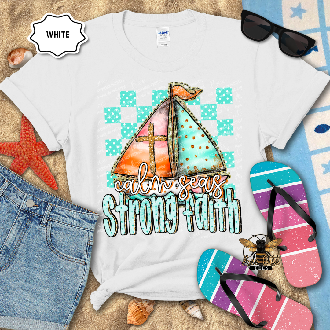White t-shirt with sailboat design and 'calm seas strong faith' text, surrounded by beach accessories.