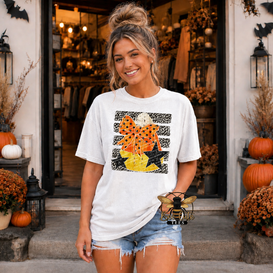 A woman stands in front of a storefront doorway wearing a white graphic T-shirt with a Halloween-themed print featuring a black leopard print bow, orange polka-dot details, a yellow pumpkin, and black stars. She is also wearing denim shorts.
