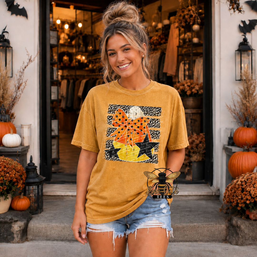 A woman stands in front of a boutique doorway wearing a mustard yellow graphic T-shirt with a Halloween-themed bow, stars, and pumpkin design, paired with light blue denim shorts.