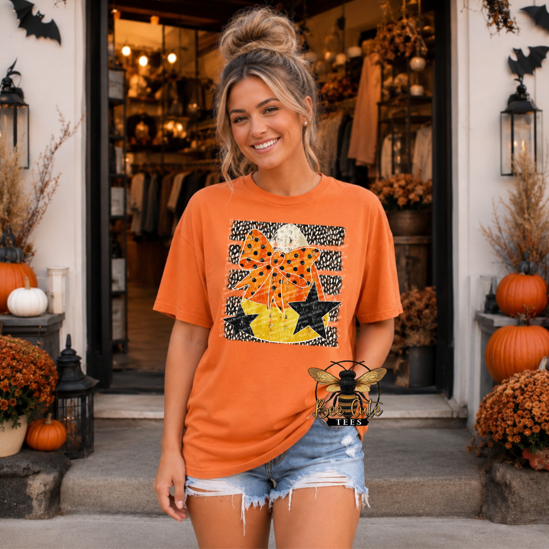 A woman stands in front of a boutique doorway wearing an orange graphic T-shirt with a Halloween-themed print featuring stars, bats, pumpkins, and a bee, paired with denim shorts.