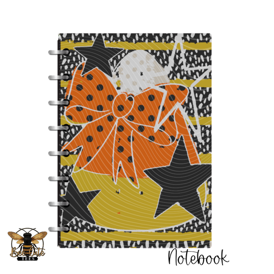 A spiral-bound notebook with a bold graphic cover featuring an orange bow, black stars, and yellow stripes on a black-and-white patterned background, shown on a white backdrop with a bee logo and the word 'Notebook' at the bottom.