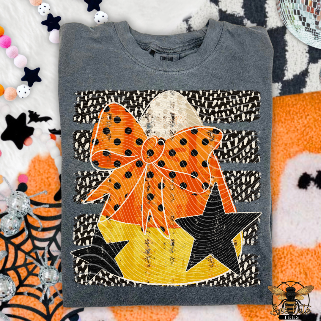 A folded gray crewneck sweatshirt featuring a large Halloween-themed graphic print with an orange polka-dot bow, a yellow striped star, and black-and-white patterned stripes, styled on a festive background with candy, stars, and spiderwebs.
