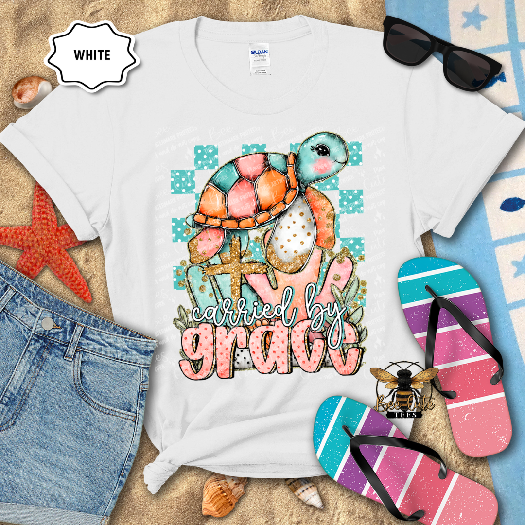 White t-shirt with a colorful turtle graphic and 'carried by grace' text, surrounded by beach items.