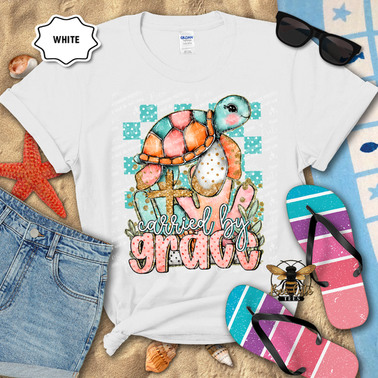 White t-shirt with a colorful turtle graphic and 'carried by grace' text, surrounded by beach items.