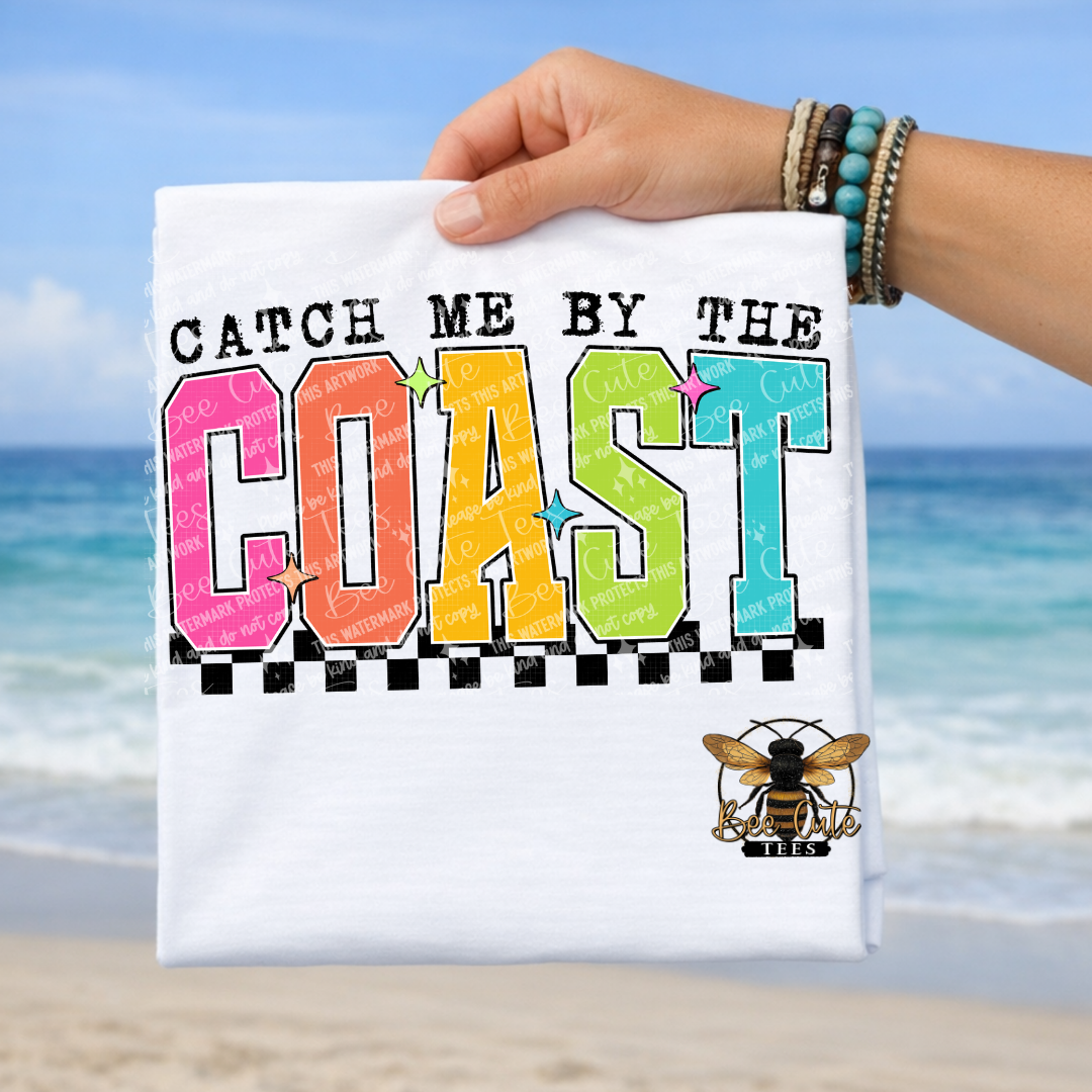 Hand holding a t-shirt with 'Catch Me By The Coast' text against a beach background