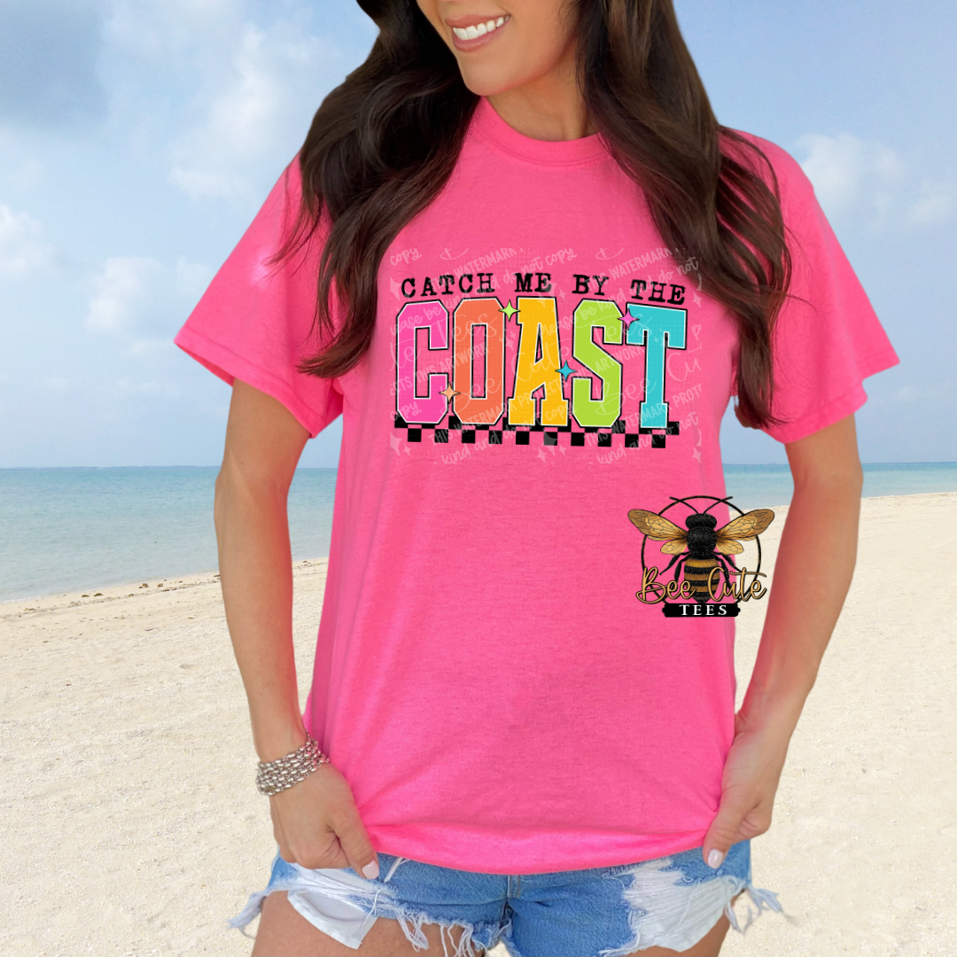 Person wearing a pink t-shirt with 'Catch Me by the Coast' design on a beach.