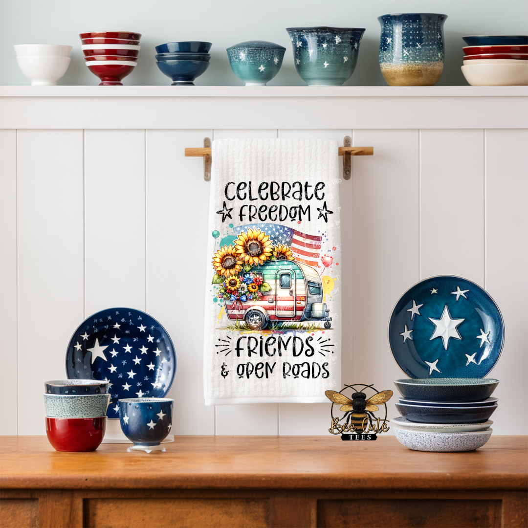 White kitchen towel hanging on a wall hook, featuring a colorful design of a vintage camper with sunflowers and an American flag, along with the phrase “Celebrate Freedom, Friends & Open Roads,” displayed in a styled kitchen with patriotic dishes and bowls.
