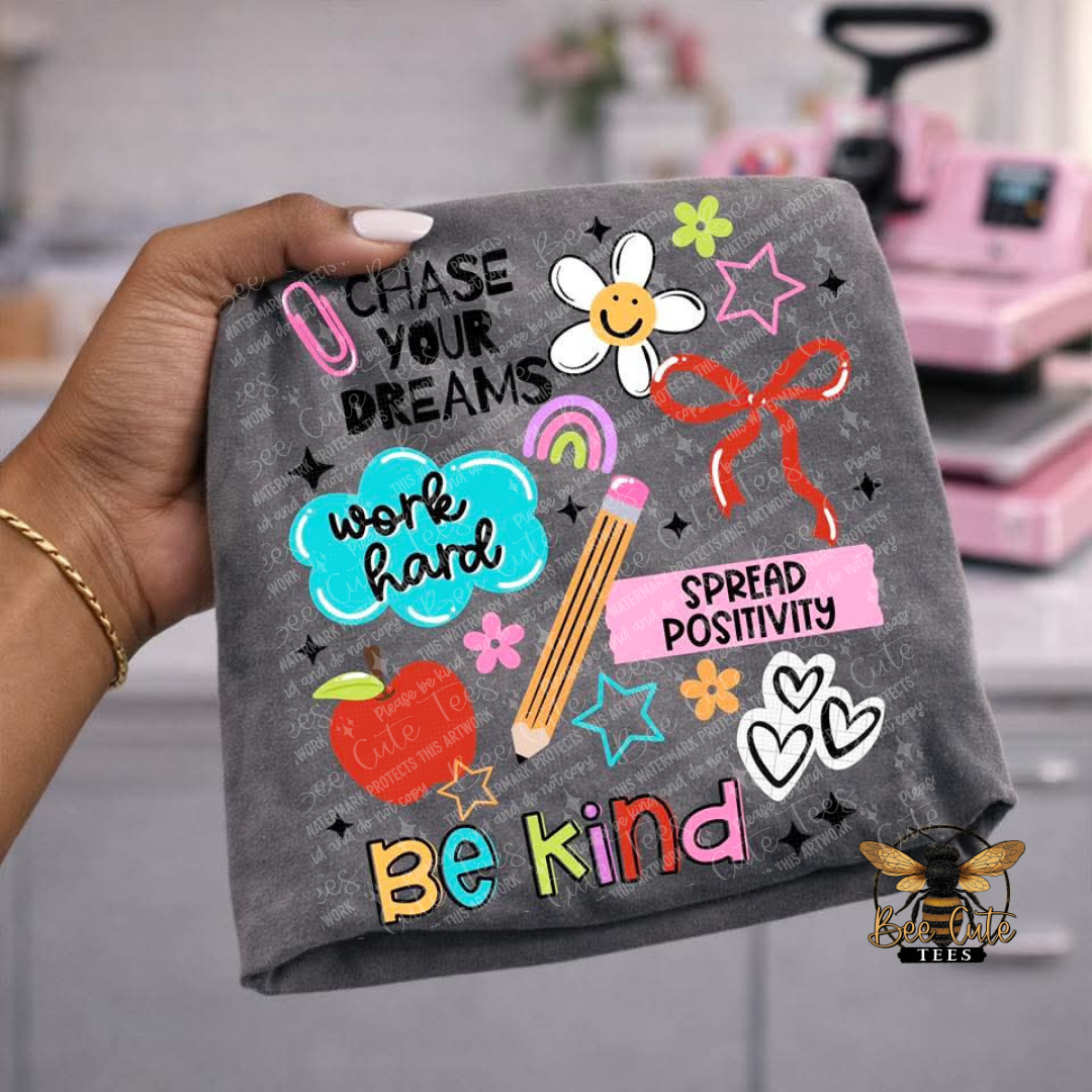 A hand holds a dark gray t-shirt featuring colorful school-themed graphics and motivational text including 'Chase Your Dreams', 'Work Hard', 'Spread Positivity', and 'Be Kind'. The background shows a blurred pink classroom setting with a chalkboard and chalk.