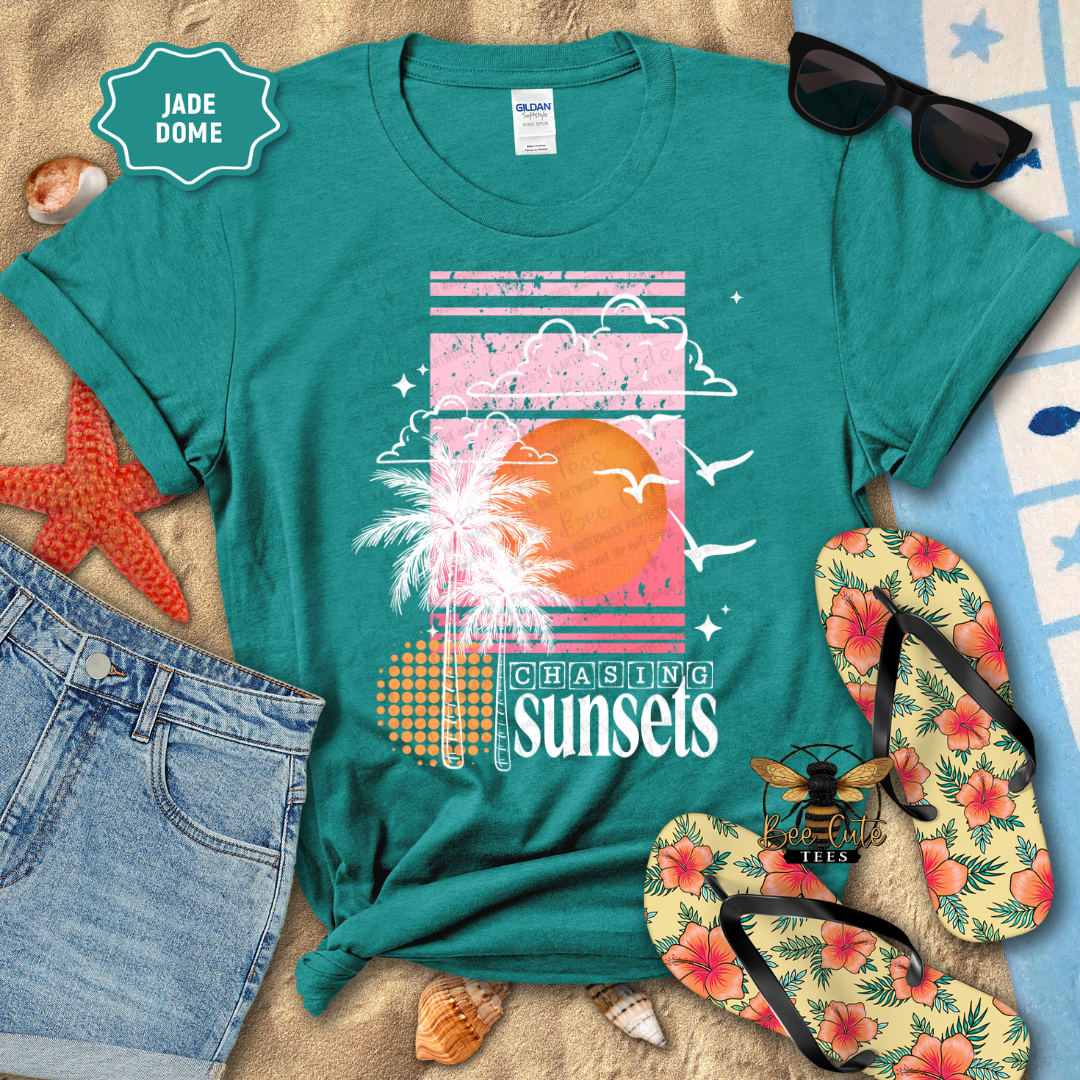 Teal t-shirt with 'Chasing Sunsets' design on a beach-themed background with sunglasses, flip-flops, and denim shorts.