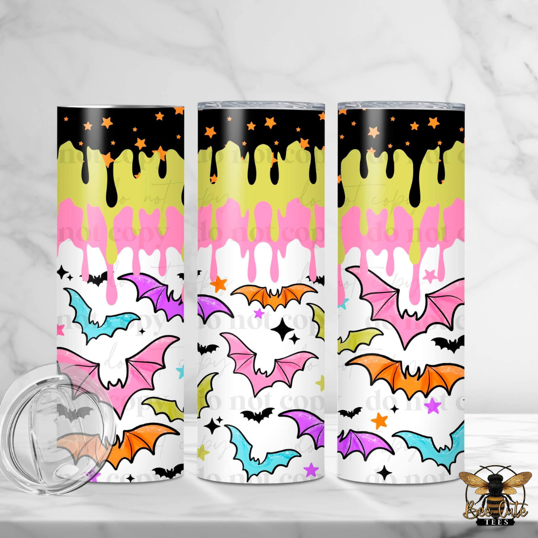 Three slim tumblers with a colorful Halloween-themed drip design, featuring black, yellow, pink, purple, orange, and blue bats on a white background, shown on a marble surface with a clear lid in the foreground.