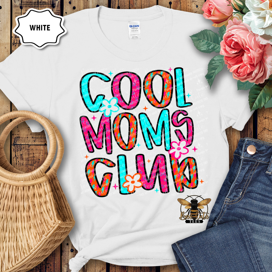 White t-shirt with colorful 'Cool Moms Club' text and flowers on a wooden surface