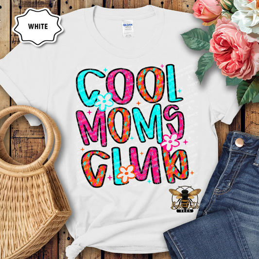 White t-shirt with colorful 'Cool Moms Club' text and flowers on a wooden surface