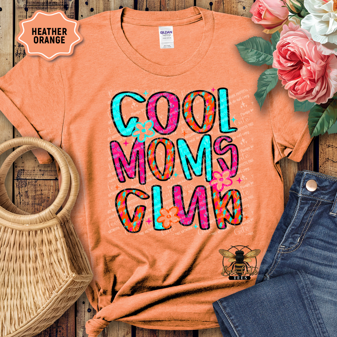 Orange t-shirt with 'Cool Moms Club' text, placed on a wooden surface with flowers and denim jeans.