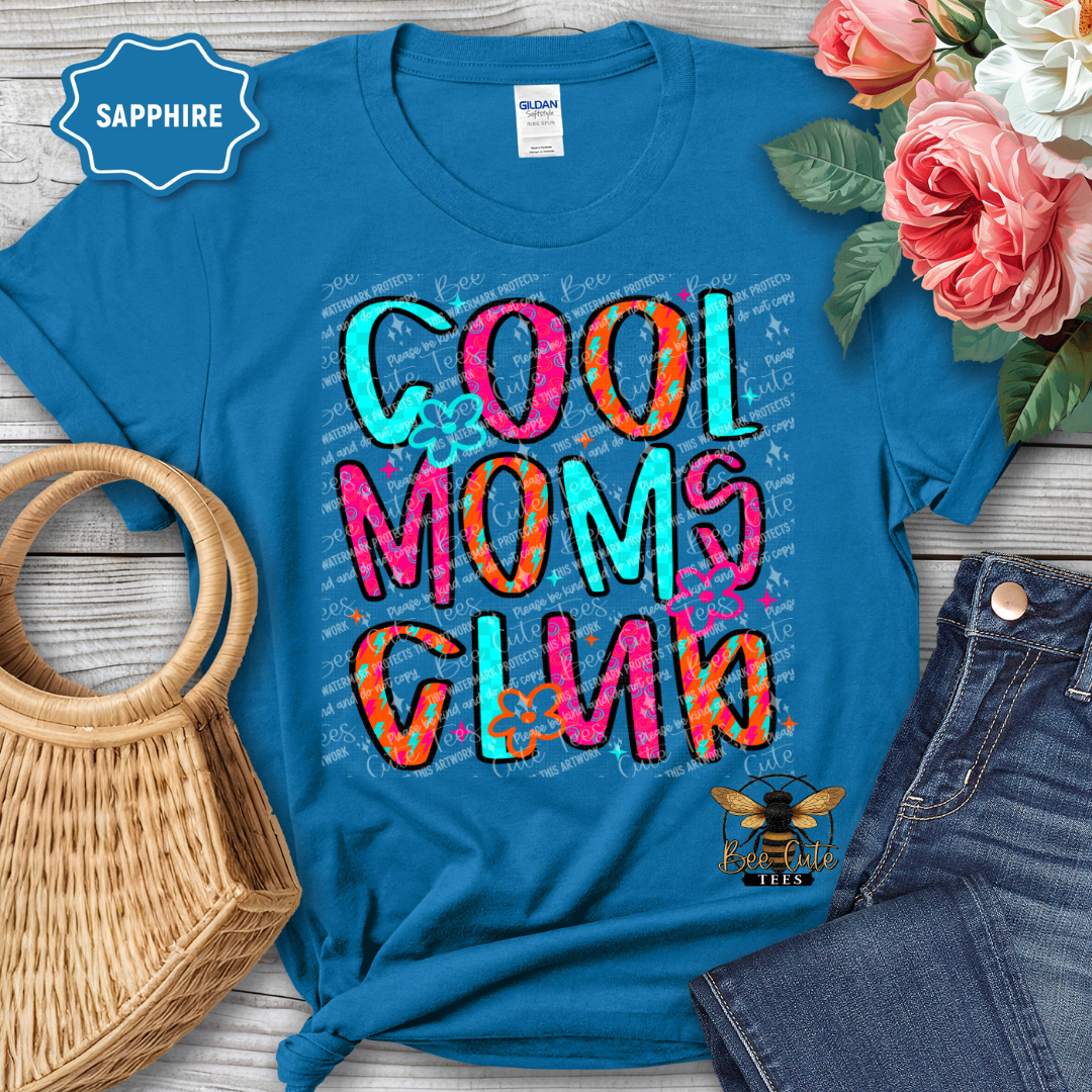 Blue t-shirt with colorful 'Cool Moms Club' text, surrounded by flowers and denim jeans on a wooden surface.