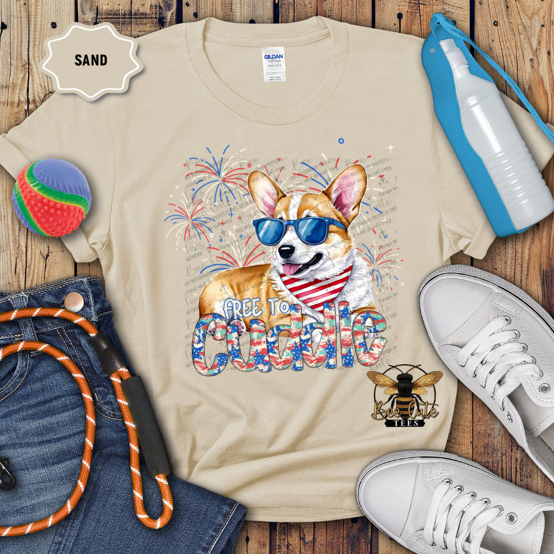 A beige short-sleeved t-shirt with a graphic print of a corgi wearing sunglasses and the text 'FREE TO Cuddle', surrounded by a patriotic red, white, and blue design, accompanied by a depiction of a beach ball, rope, and sunglasses.