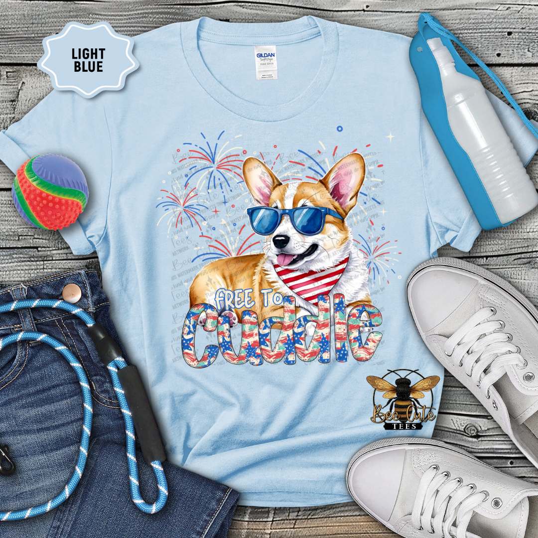 Light blue t-shirt with a dog graphic and 'Free to Cuddle' text, surrounded by jeans, shoes, and accessories on a wooden surface.