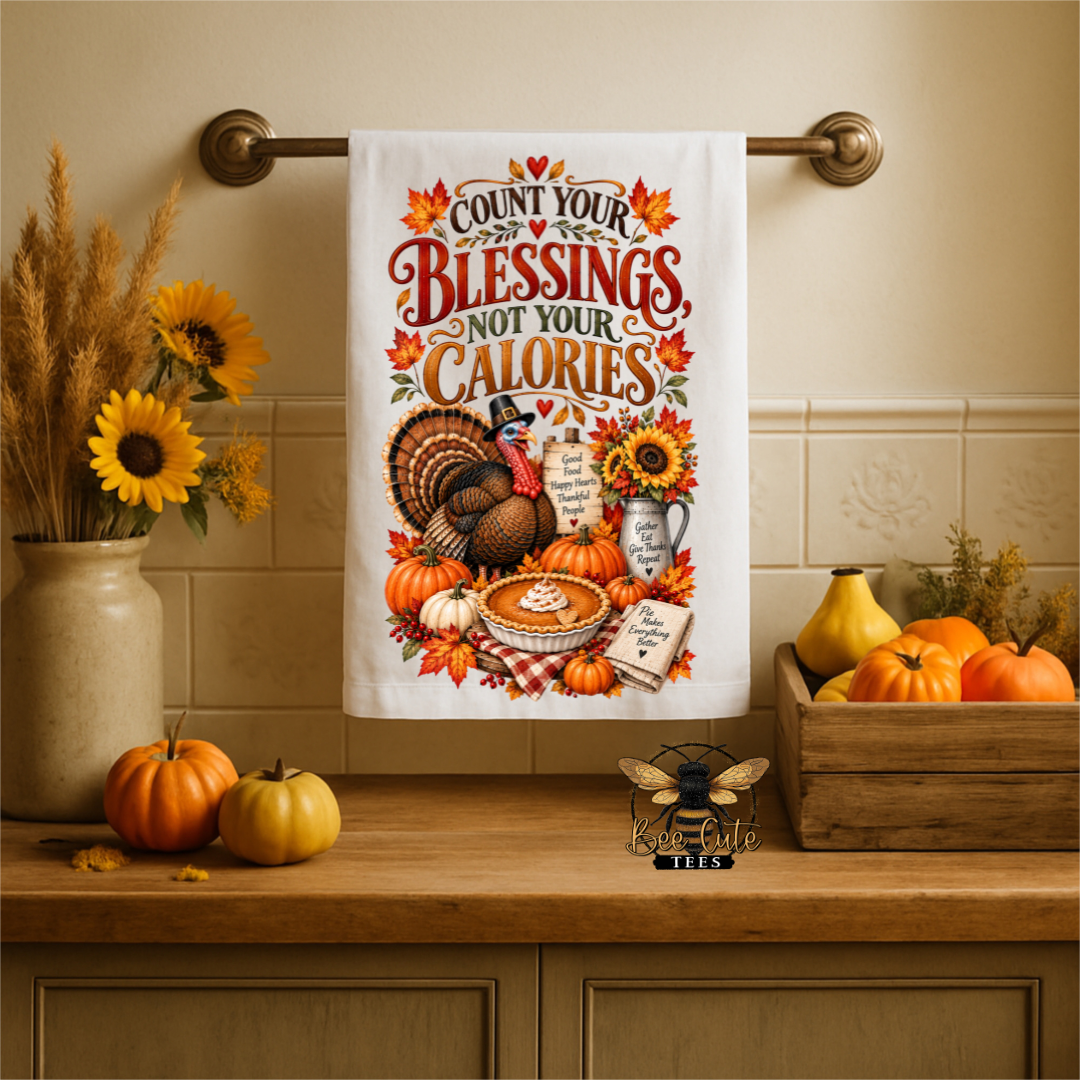 A white kitchen towel with a colorful Thanksgiving-themed illustration and the phrase 'COUNT YOUR BLESSINGS NOT YOUR CALORIES' hangs on a brass towel bar above a wooden counter decorated with pumpkins, sunflowers, and a small bee figurine.