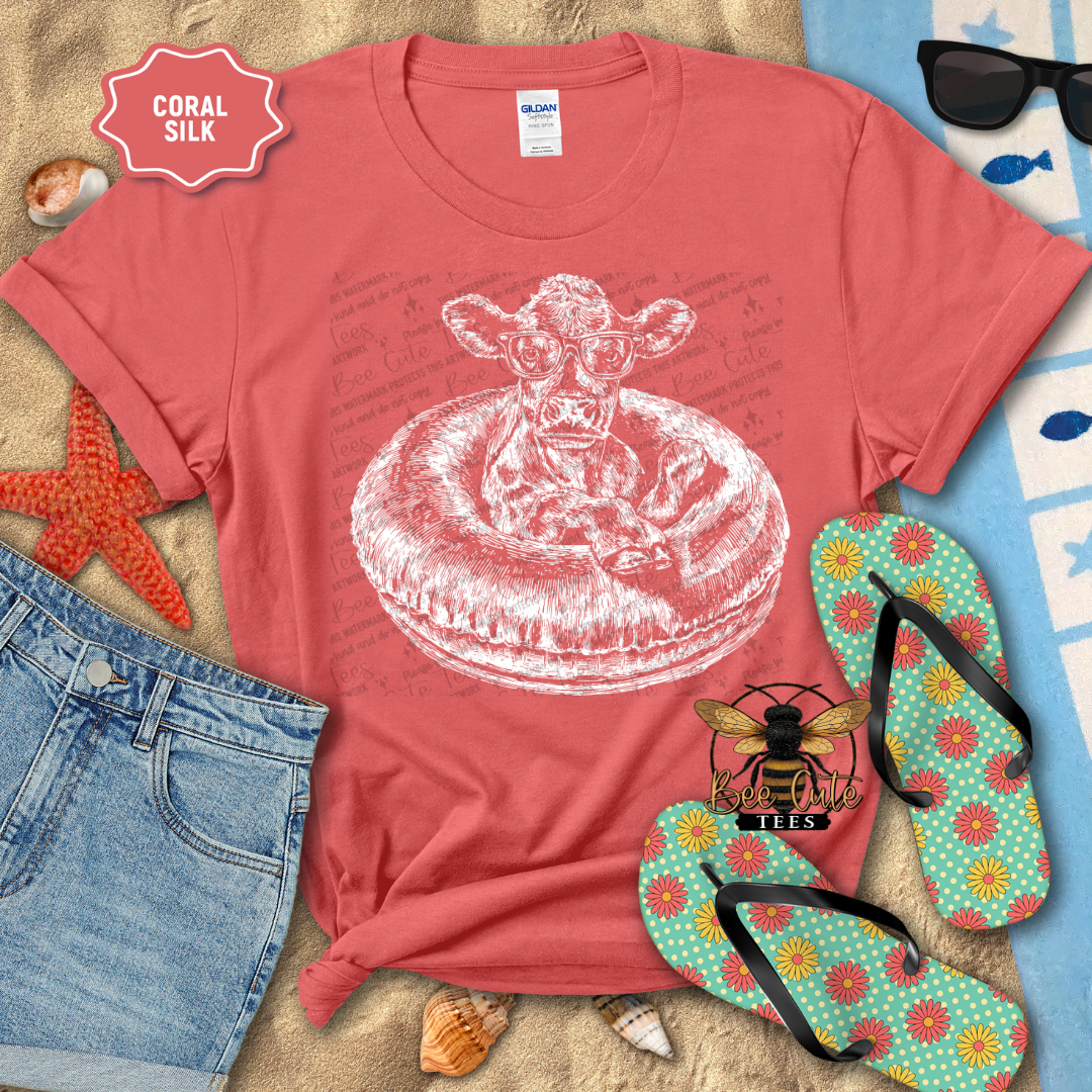 Coral t-shirt with a cow on an inner tube graphic, surrounded by beach items.