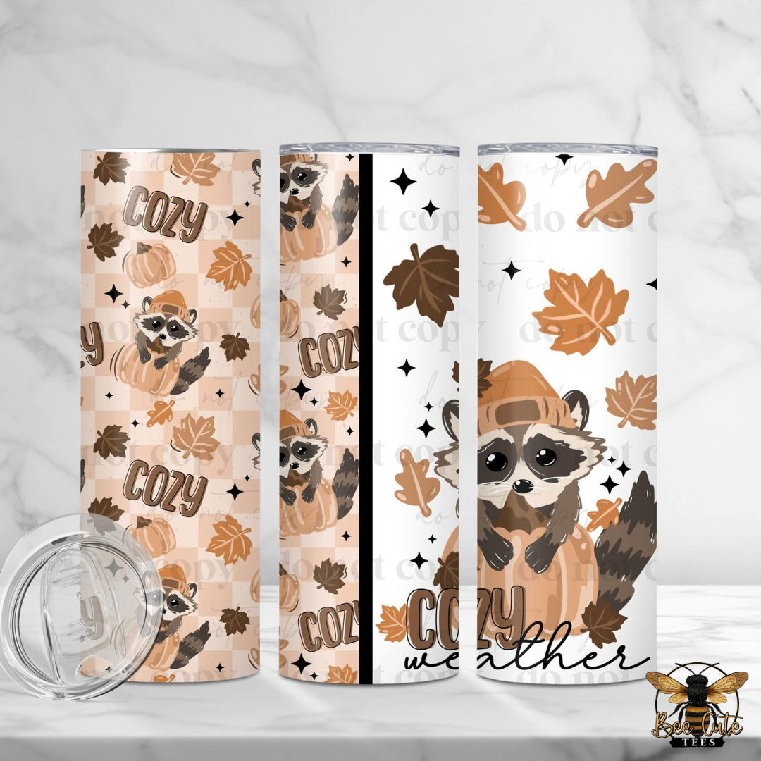 Three views of a tall insulated tumbler with a fall raccoon and autumn leaf pattern, shown on a light marble surface with a clear lid in front. The designs feature tan, brown, and beige tones with the words 'Cozy weather' and illustrated raccoons wearing hats.