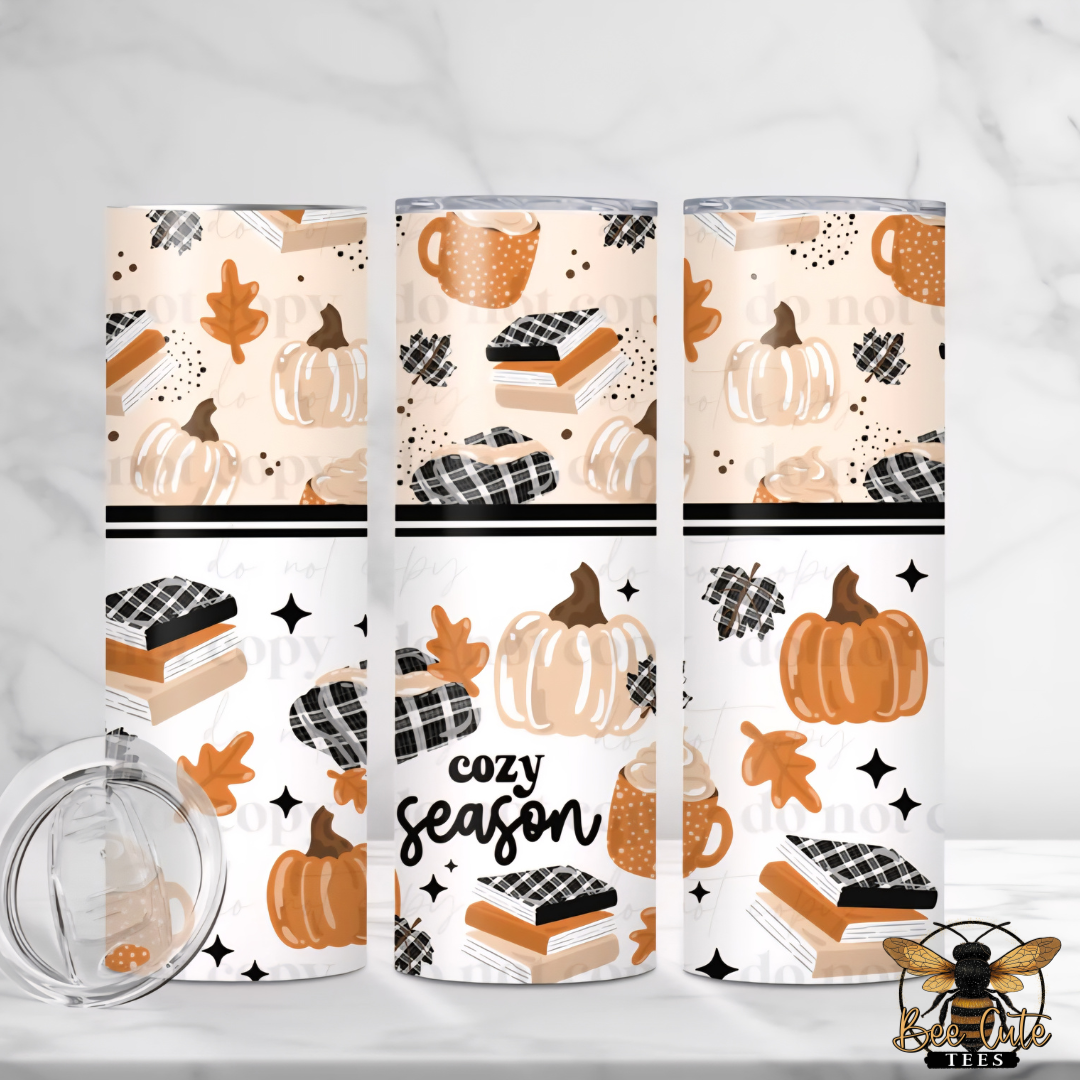 Three tall insulated tumblers with clear lids are shown on a light marble-like surface. The tumblers have an autumn-themed wrap design with pumpkins, leaves, books, coffee cups, stars, and the words 'cozy season.