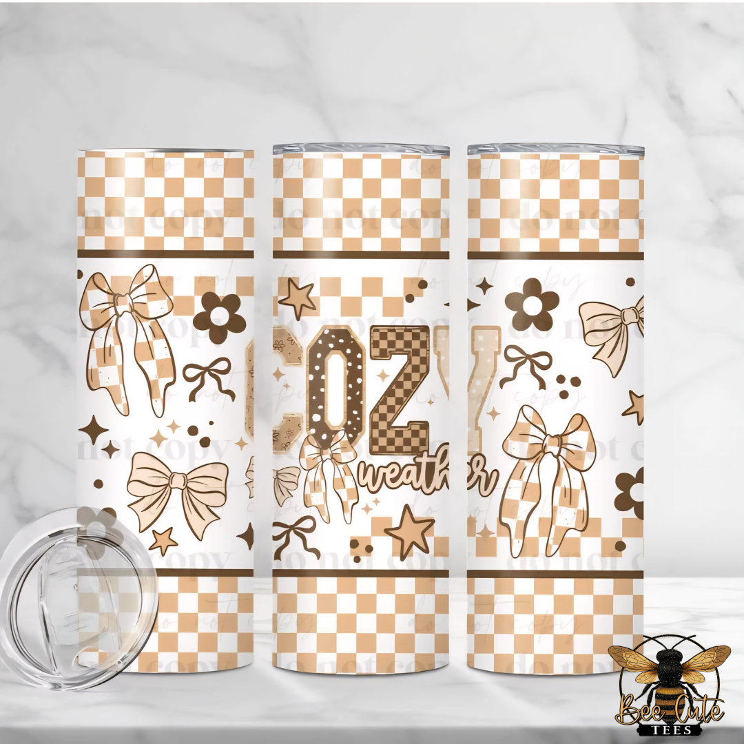 Three tall insulated tumblers with clear lids are shown on a light marble surface and backdrop. The tumblers have a white and tan checkerboard pattern with bows, stars, flowers, and the word cozy.