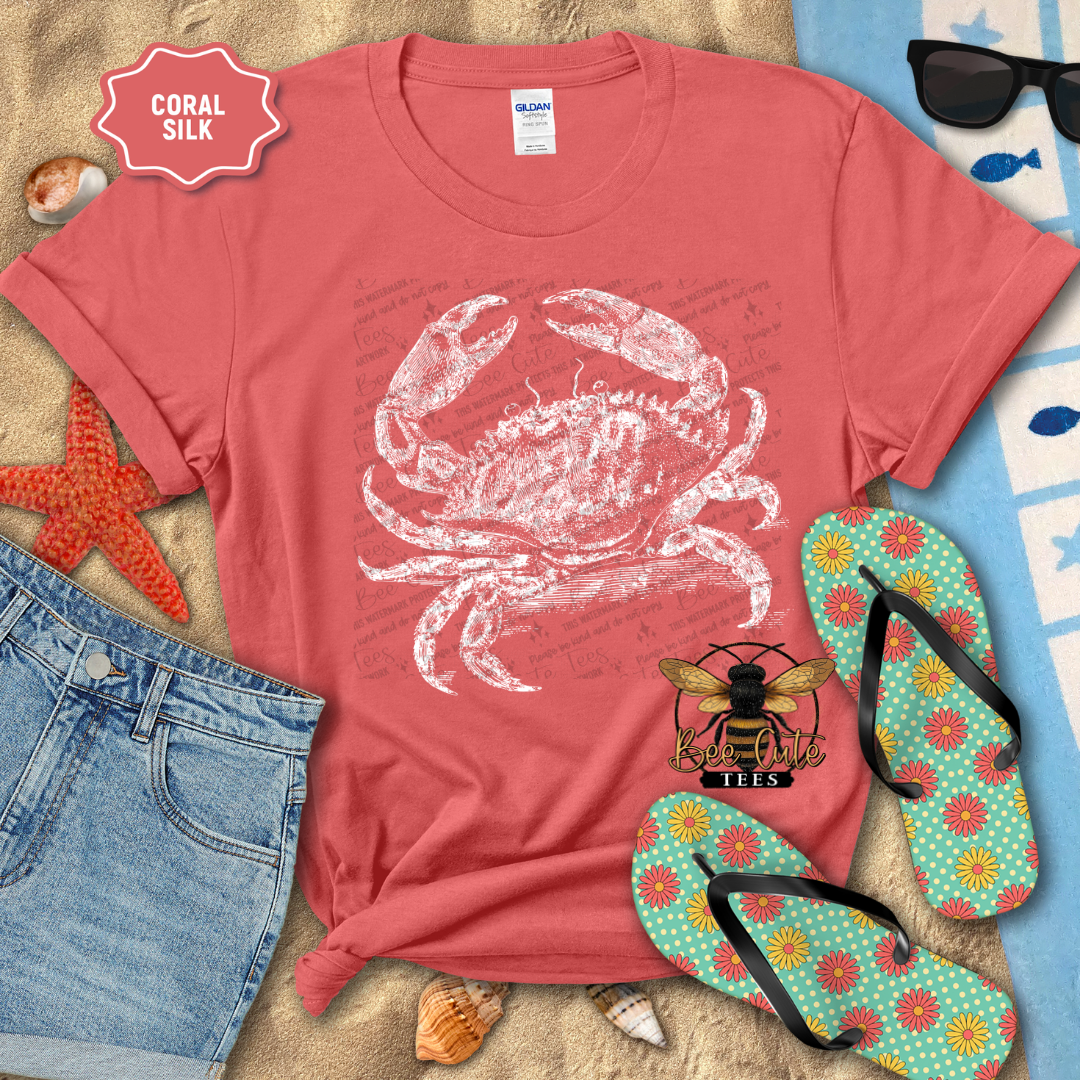 Coral t-shirt with a white crab design, surrounded by beach accessories on sand.